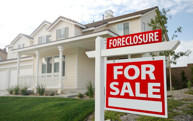 Photo of Foreclosure For Sale sign in front of beige house and green yard. 