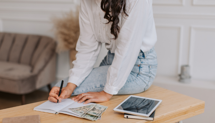 Woman sitting on edge of table budgeting with tablet, notebook, and cash. 