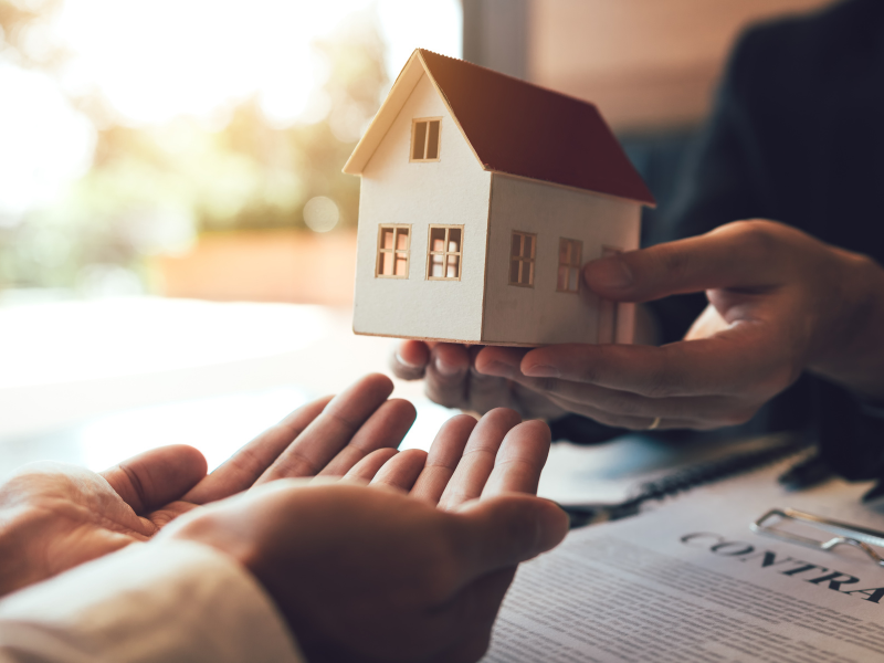 Open hands passing model house across table over contract paperwork.