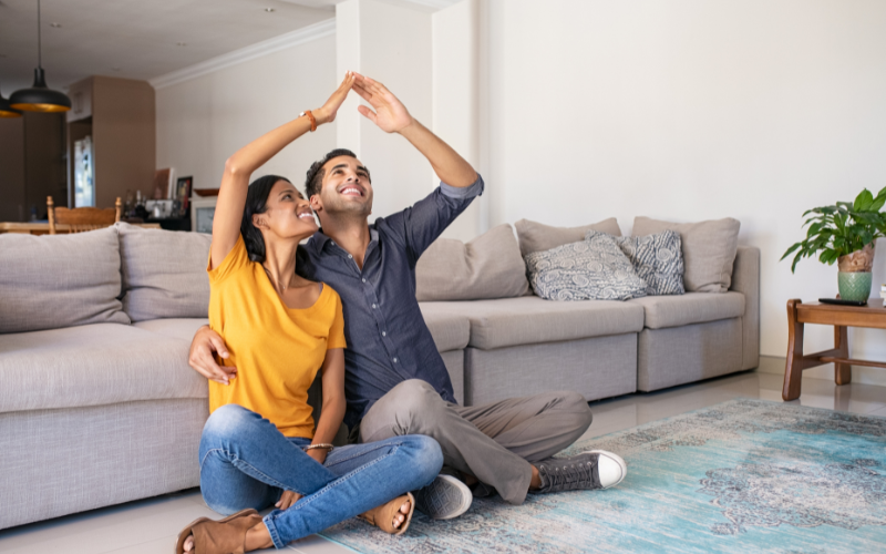 Man and woman living room holding arms above head to look like a house above their heads. 